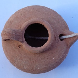 May include: A terracotta oil lamp with a small handle and a wick hole. The lamp is made of a reddish-brown clay and has a simple, rustic design.