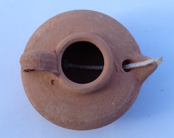 Könnte beinhalten: Eine terracotta-Öllampe mit einem kleinen Griff und einem Dochtloch. Die Lampe ist aus rotbraunem Ton gefertigt und hat ein schlichtes, rustikales Design.