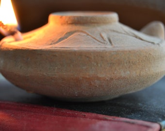 Könnte beinhalten: Eine terracotta-Öllampe mit einer brennenden Flamme im Inneren. Die Lampe hat ein schlichtes Design mit einem runden Korpus und einem Ausguss für den Docht. Die Lampe steht auf einer rot-schwarzen Oberfläche.