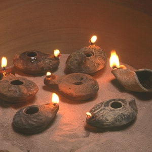 Może przedstawiać: Siedem małych, glinianych lamp naftowych z zapłonionymi knotami, ułożonych w kółko na piaszczystym podłożu.