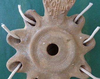 Puede incluir: Una lámpara de aceite de terracota con siete mechas, cada una con un cordón de algodón blanco. La lámpara tiene una abertura circular en el centro y un diseño en relieve en la parte superior con un árbol y una inscripción.