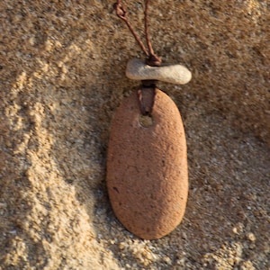 May include: A brown stone pendant with a hole in the centre, hanging from a brown cord. The pendant is attached to a smaller, light brown stone.