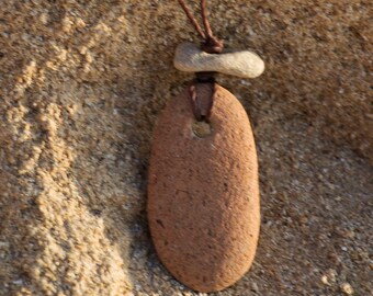 May include: A brown stone pendant with a hole in the center, hanging from a brown cord. The pendant is attached to a smaller, light brown stone.