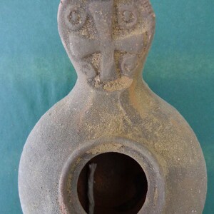 May include: A gray clay oil lamp with a cross design on the top. The lamp has a round opening for a wick.