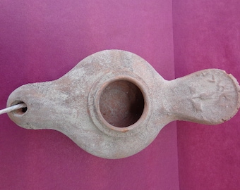 May include: A terracotta oil lamp with a round opening and a long spout. The lamp has a rough, textured surface and is slightly dusty.