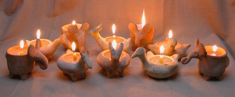 Puede incluir: Una colecci&oacute;n de nueve candelabros de cer&aacute;mica con forma de animales, incluyendo un elefante, un p&aacute;jaro, un ciervo y una tortuga. Cada candelabro tiene una vela peque&ntilde;a en su interior, y las velas est&aacute;n encendidas, proyectando un c&aacute;lido resplandor.