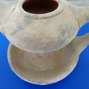 May include: A terracotta oil lamp with a spout and a separate base. The lamp is made of unglazed clay and has a rustic, aged appearance.