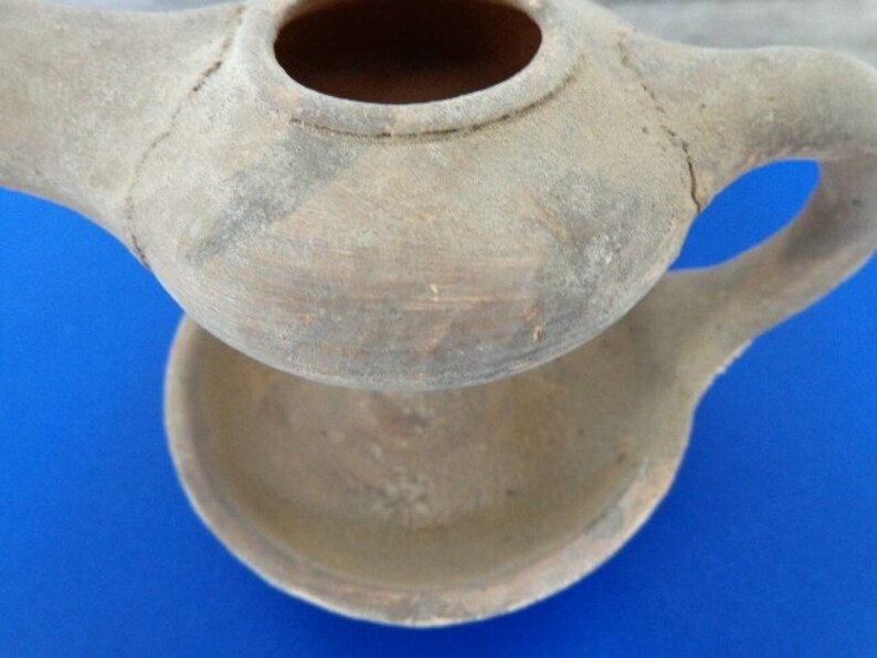 May include: A terracotta oil lamp with a spout and a separate base. The lamp is made of unglazed clay and has a rustic, aged appearance.