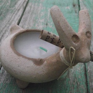 Puede incluir: Un portavelas de cer&aacute;mica con forma de animal estilizado con una vela blanca en su interior. El portavelas es de color marr&oacute;n y blanco y est&aacute; decorado con una cinta marr&oacute;n y un peque&ntilde;o trozo de papel con texto.