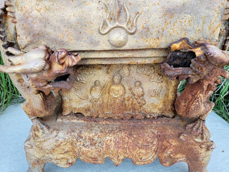 May include: A vintage cast iron incense burner with a dragon design. The burner has a rectangular base with four legs and a raised top with a dragon head on each side. The top of the burner has a relief design of a Buddha figure surrounded by two figures. The burner is rusty and shows signs of wear.