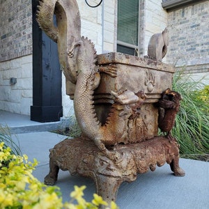 May include: A rusty metal planter with a dragon design. The planter is on a stand with four legs. The planter is in front of a brick wall.