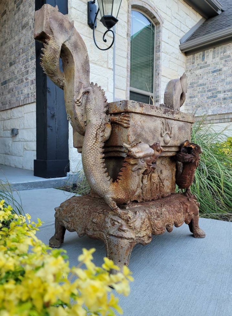 May include: A rusty metal planter with a dragon design. The planter is on a stand with four legs. The planter is in front of a brick wall.