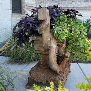 May include: A rusty metal dragon planter with a leafy green plant and a purple plant growing inside. The planter is sitting on a concrete patio.