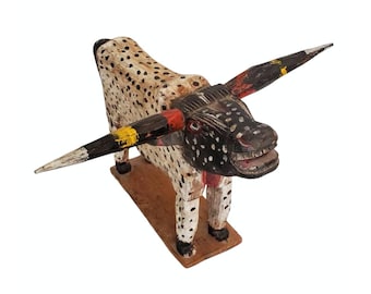 May include: A hand-carved wooden bull figurine with black and white spots, red and yellow painted horns, and a toothy grin. The bull is standing on a wooden base.