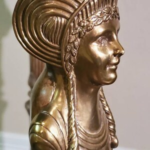 May include: Close-up of a gold-toned sculpture of a woman's bust, part of a table leg. The woman has a detailed headdress and braided hair. The sculpture is made of a metallic material.