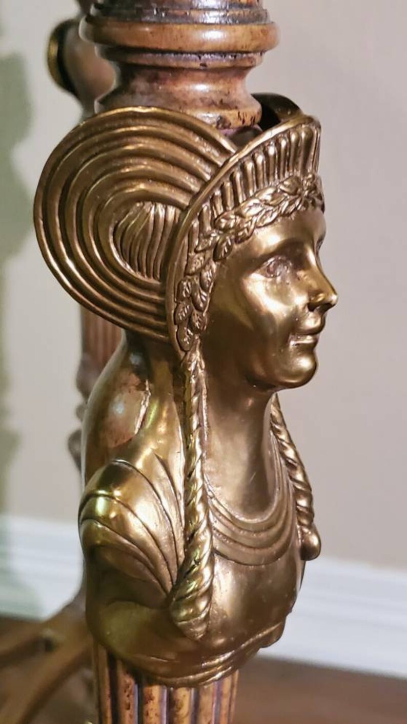 May include: Close-up of a gold-toned sculpture of a woman's bust, part of a table leg. The woman has a detailed headdress and braided hair. The sculpture is made of a metallic material.