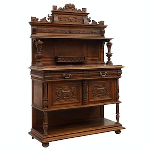 May include: A large, intricately carved wooden sideboard with a marble top. The sideboard has two doors with ornate carvings and a shelf below. The top section has a shelf and is decorated with a carved crest.