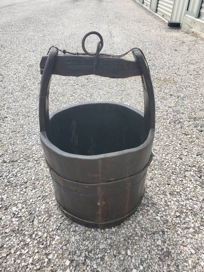 Puede incluir: Un cubo de pozo de madera marr&oacute;n oscuro con un anillo y un asa de metal. El cubo est&aacute; desgastado y tiene un aspecto r&uacute;stico.