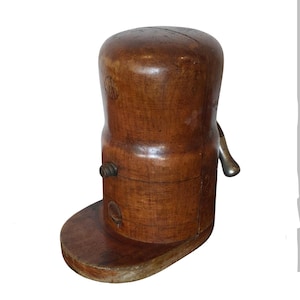 May include: A vintage wooden hat stretcher with a dark brown finish. The hat stretcher has a cylindrical body, a base, and a metal handle. The wood shows signs of age and wear, adding to its antique charm. The stretcher is designed to maintain or reshape hats.