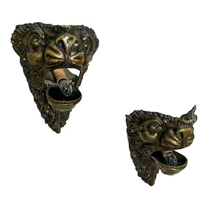 May include: Two antique bronze lion head wall sconces with a decorative design. Each sconce has a small bowl for holding a candle or other light source.