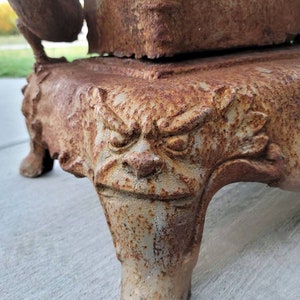 May include: A rusty cast iron wood-burning stove with a decorative base featuring a grotesque face. The stove has a large opening for burning wood and a smaller opening for ash removal.