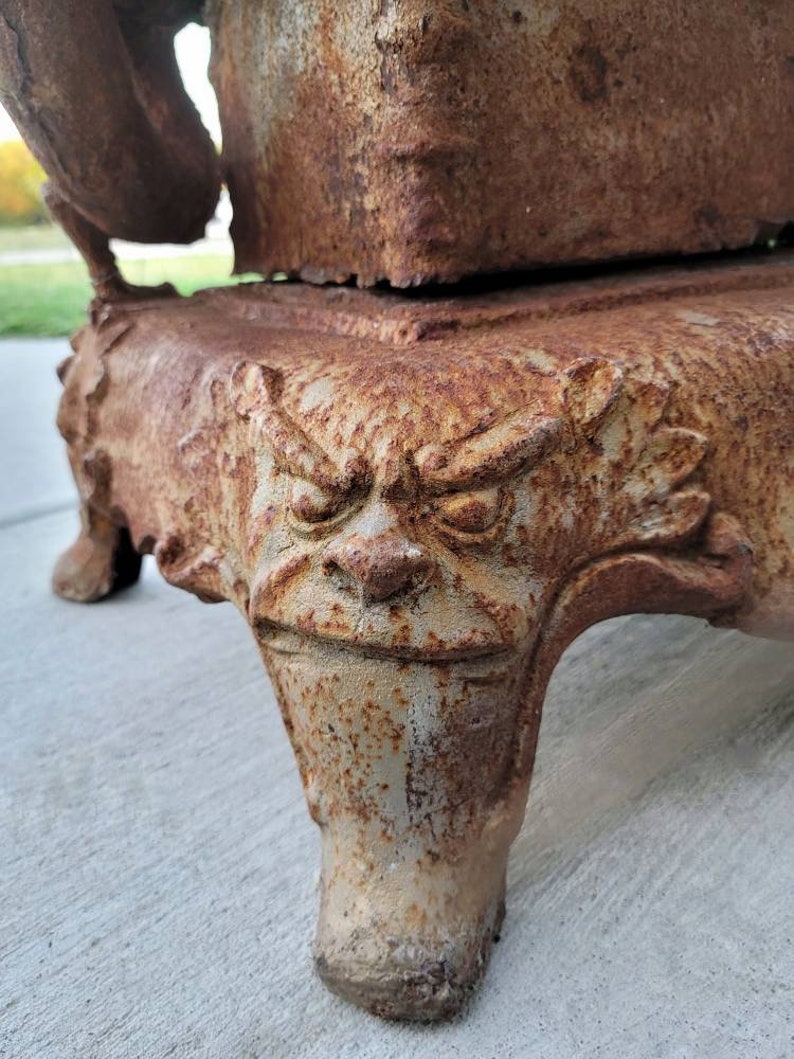 May include: A rusty cast iron wood-burning stove with a decorative base featuring a grotesque face. The stove has a large opening for burning wood and a smaller opening for ash removal.