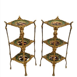 May include: A pair of gold metal three-tiered stands with green and gold decorative plates. Each plate features a painted scene of people in a garden setting.