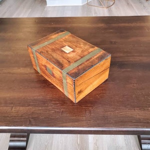 May include: A wooden box with brass accents sits on a dark wood table. The box has a light wood top and is closed.