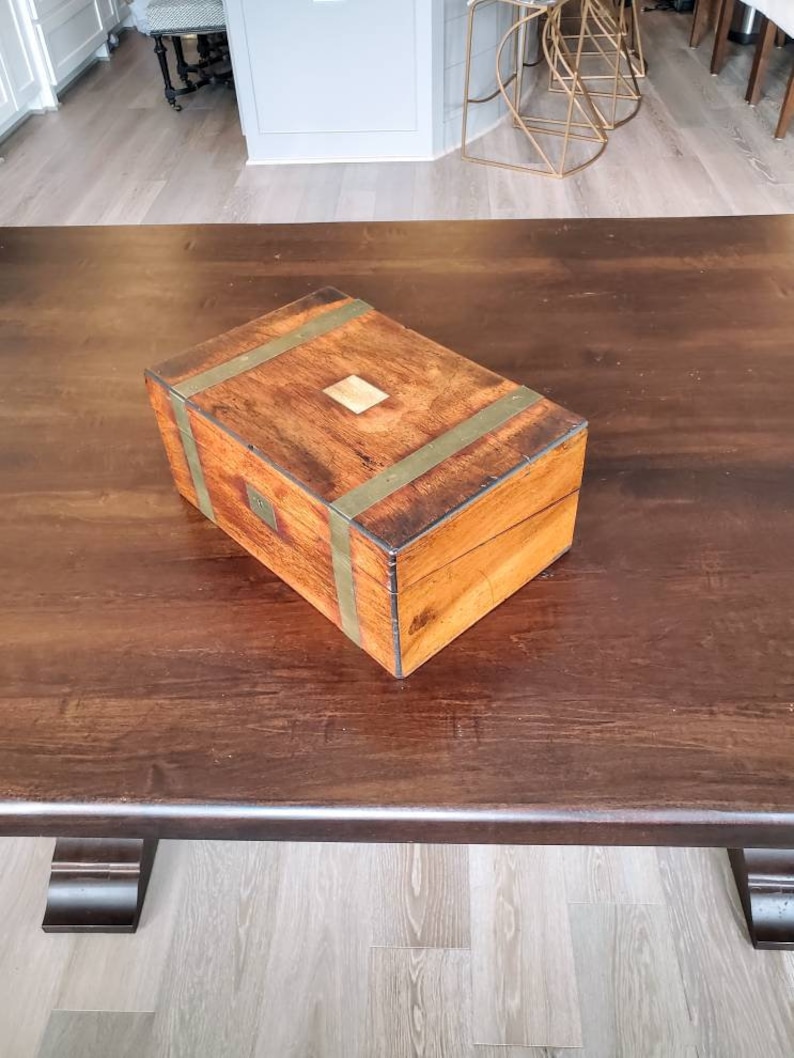 May include: A wooden box with brass accents sits on a dark wood table. The box has a light wood top and is closed.