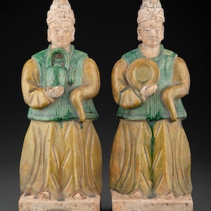 May include: Two ceramic figurines of people wearing green and yellow robes and tall hats. The figurines are standing on square bases.