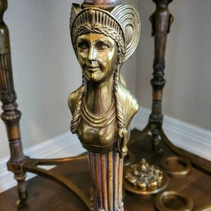 May include: Ornate brass table with a dark top. The table features a detailed bust of a woman with braided hair and a decorative headpiece. The table's legs and supports are also intricately designed, showcasing a vintage aesthetic.