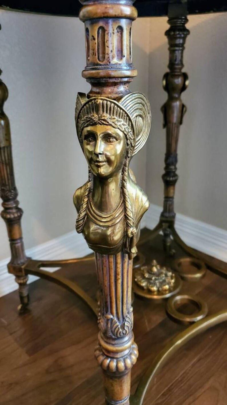 May include: Ornate brass table with a dark top. The table features a detailed bust of a woman with braided hair and a decorative headpiece. The table's legs and supports are also intricately designed, showcasing a vintage aesthetic.