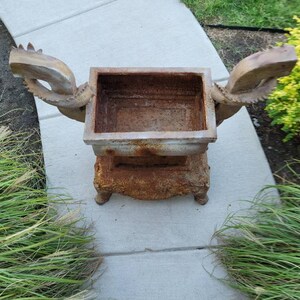 May include: A rusty metal planter with two dragon-shaped handles. The planter is on a concrete surface.
