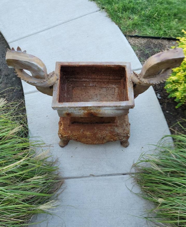 May include: A rusty metal planter with two dragon-shaped handles. The planter is on a concrete surface.
