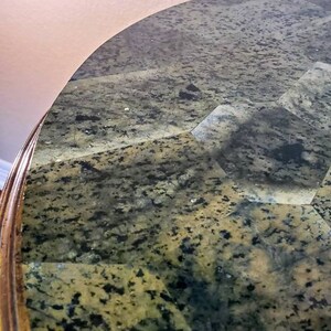 May include: Close-up of a table with a patterned granite top. The surface is a mix of green, black, and gray, with a polished finish. The table's edge is a warm, golden brown.