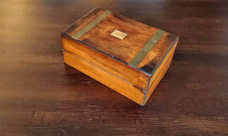 May include: A wooden box with brass accents. The box has a hinged lid and a brass plate on the top.
