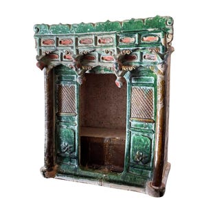 May include: A detailed, antique ceramic structure with a green and brown color scheme. The piece features intricate carvings, including floral and geometric patterns, and a small interior space. The top has a decorative roof-like design with red accents.