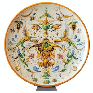 May include: A large, round, white ceramic plate with an orange rim. The plate features a detailed, symmetrical design of stylized birds, flowers, and leaves in shades of yellow, green, blue, and pink.