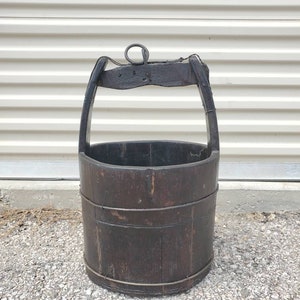 Puede incluir: Un cubo de pozo de madera vintage con un asa de metal y una cuerda atada. El cubo es de color marr&oacute;n oscuro y tiene un aspecto desgastado y r&uacute;stico.