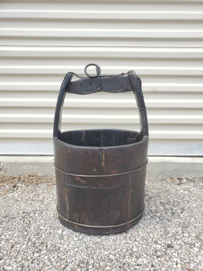 Puede incluir: Un cubo de pozo de madera vintage con un asa de metal y una cuerda atada. El cubo es de color marr&oacute;n oscuro y tiene un aspecto desgastado y r&uacute;stico.
