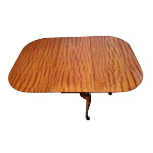 May include: A wooden oval-shaped table with a drop leaf on each side. The table has a rich brown color and a wood grain pattern. The table has four legs with a cabriole design.