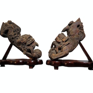 May include: A pair of intricately carved wooden sculptures of mythical creatures, each mounted on a dark wood base. The sculptures are dark brown and feature detailed carvings of faces, wings, and other decorative elements.