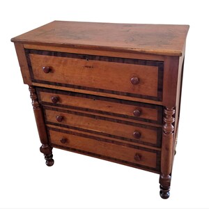 May include: A wooden chest of drawers with four drawers and turned legs. The drawers have a wood veneer with a dark brown border.