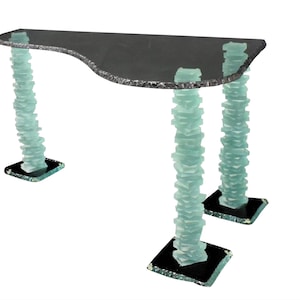 May include: A modern console table with a dark gray, curved top and three legs. The legs are constructed from stacked, light blue-green glass blocks, resting on black square bases. The table is a unique piece of furniture.
