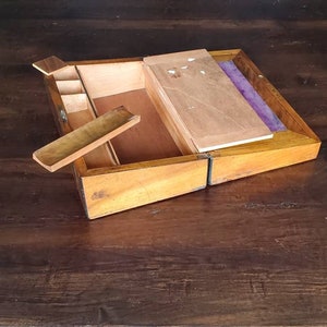 May include: A wooden writing box with a hinged lid, open to reveal a compartment with a small wooden tray and a purple felt lining. The box is made of light brown wood with a natural finish.