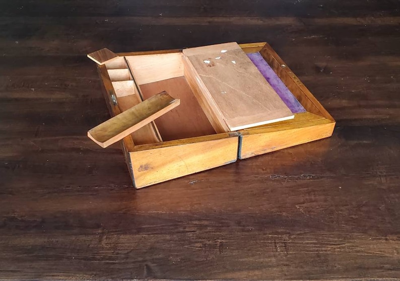 May include: A wooden writing box with a hinged lid, open to reveal a compartment with a small wooden tray and a purple felt lining. The box is made of light brown wood with a natural finish.
