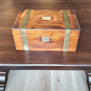 May include: A wooden box with brass accents. The box has a hinged lid and a brass lock. The box is sitting on a dark wooden surface.