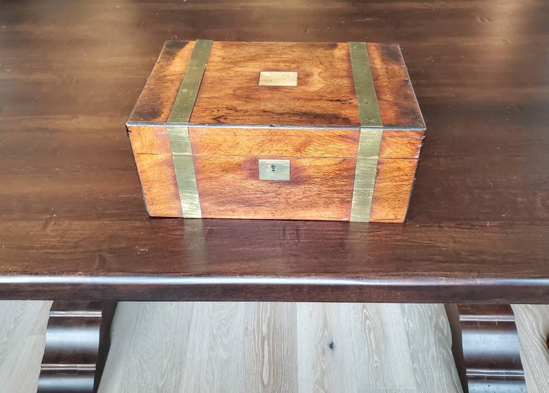 May include: A wooden box with brass accents. The box has a hinged lid and a brass lock. The box is sitting on a dark wooden surface.