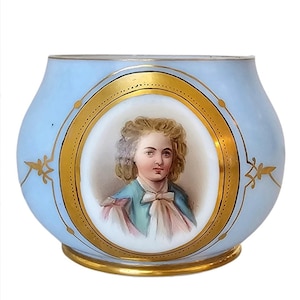 May include: A light blue glass vase with a gold rim and a painted portrait of a young person in a gold oval frame.