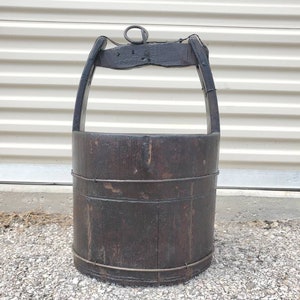 Puede incluir: Un cubo de pozo de madera vintage con un asa de metal y una cuerda atada al asa. El cubo es de color marr&oacute;n oscuro y tiene un aspecto desgastado y r&uacute;stico.